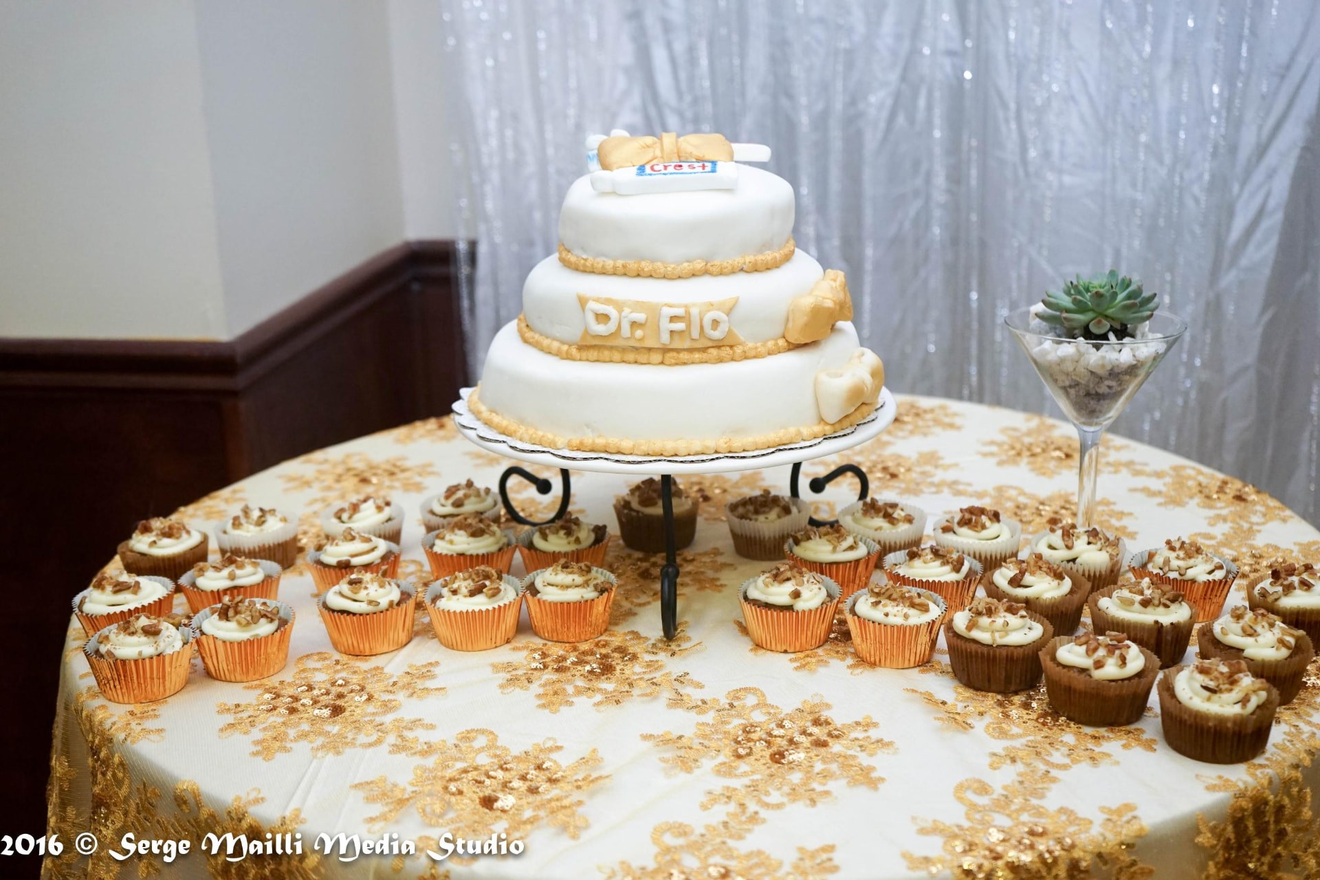 Graduation cake reading Dr. Flo surrounded by cupcakes on a gold-embroidered dessert table