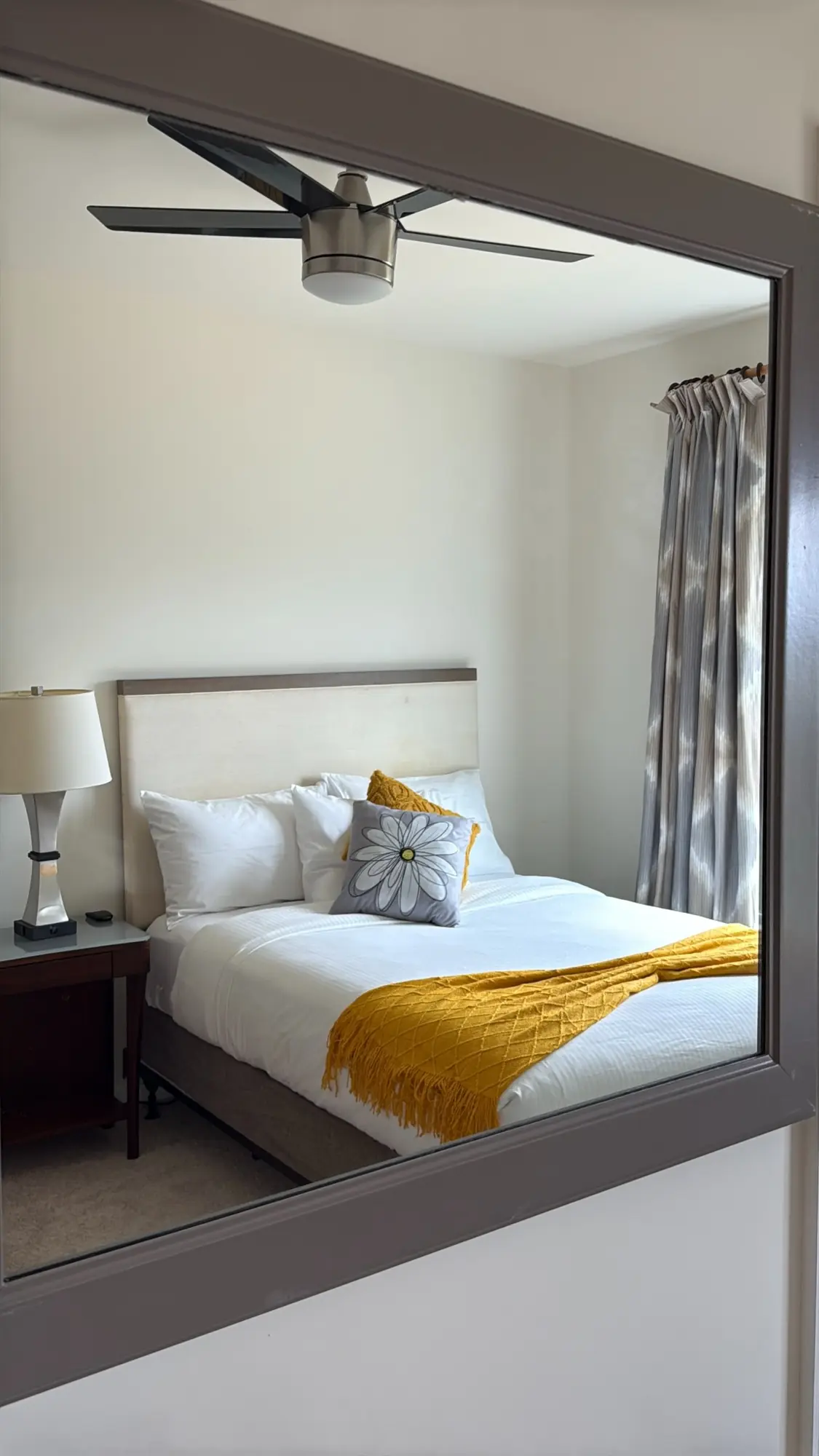 Guest bedroom with cream upholstered headboard and a gold throw, seen through a wall mirror